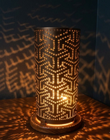 A cylindrical black table lamp with an intricate geometric laser-cut pattern, glowing with warm light from within. It sits on a wooden base, casting warm golden shadows.